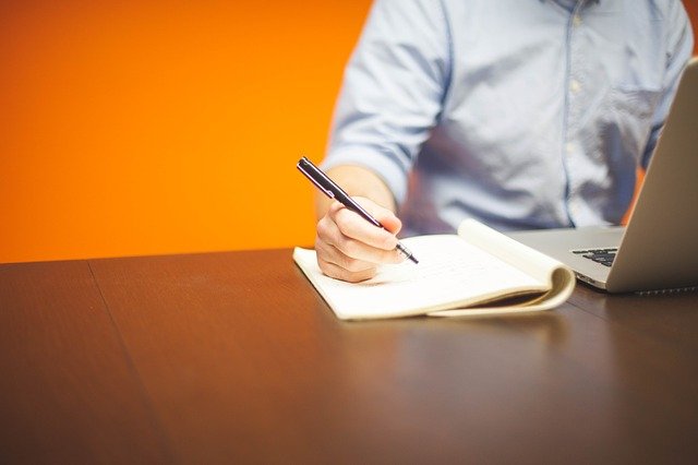 a man, reading from a laptop and taking note by hand with a notebook