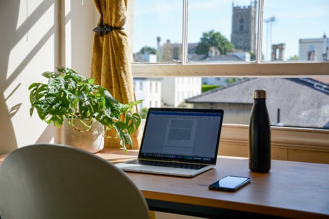 Workspace. Laptop, cell phone, and water on a desk in front of a window